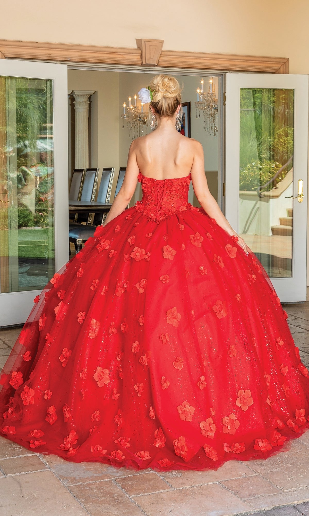 A woman in the Quinceanera Dress 1704 By Dancing Queen, featuring floral appliqués and a strapless ball gown design, stands with her back to the camera in front of open glass doors, highlighting this stunning red dress.
